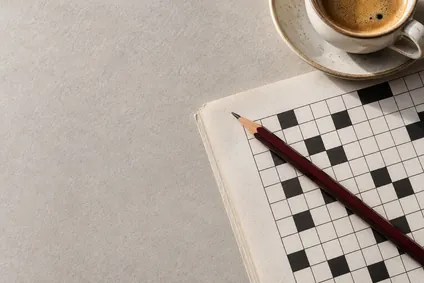 Vinröd penna och kaffekopp intill ett korsordshäfte.